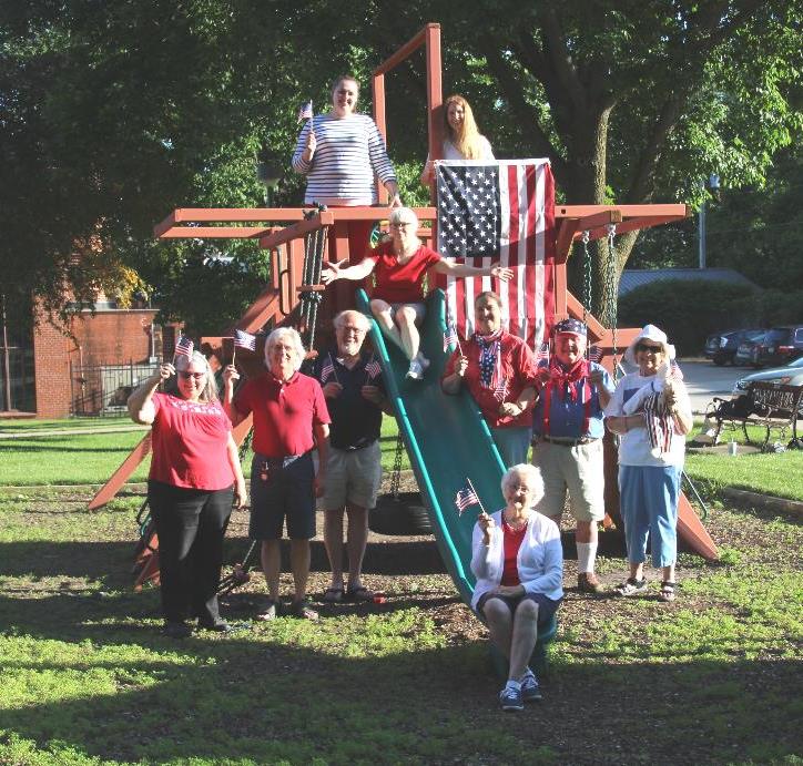 10 older people dressed in american flag style attire stand and pose for a photo on a playground set.