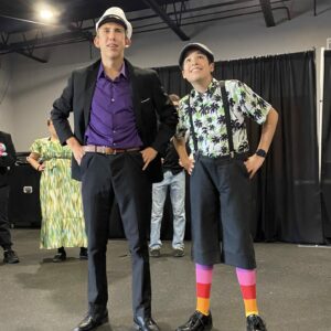Two masculine figures dressed up in costumes look off into the distance with their hands on their hips.