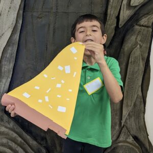 A young boy with a green shirt eats a fake piece of pizza made of paper.